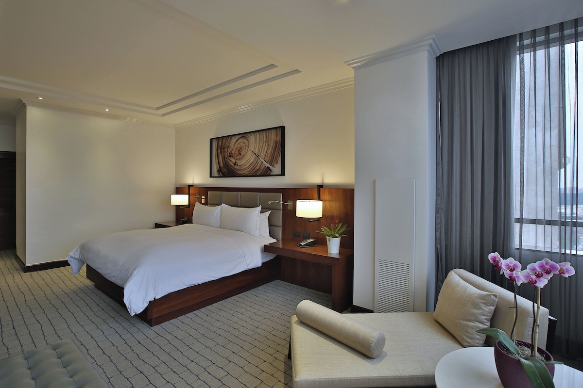 Habitación de hotel con una cama grande, lámparas de noche, un escritorio con teléfono, una planta en maceta y una ventana con cortinas.