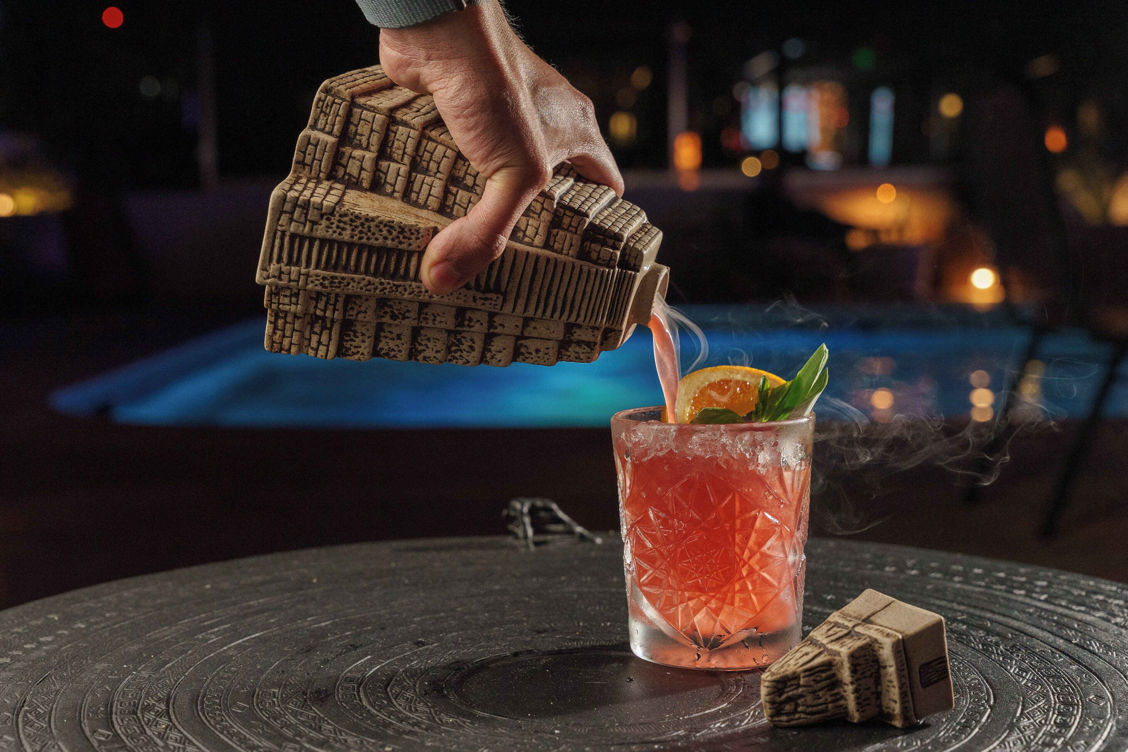 A hand pouring a drink into a glass with a straw and a slice of lime, next to a small stack of coasters on a table by a pool.