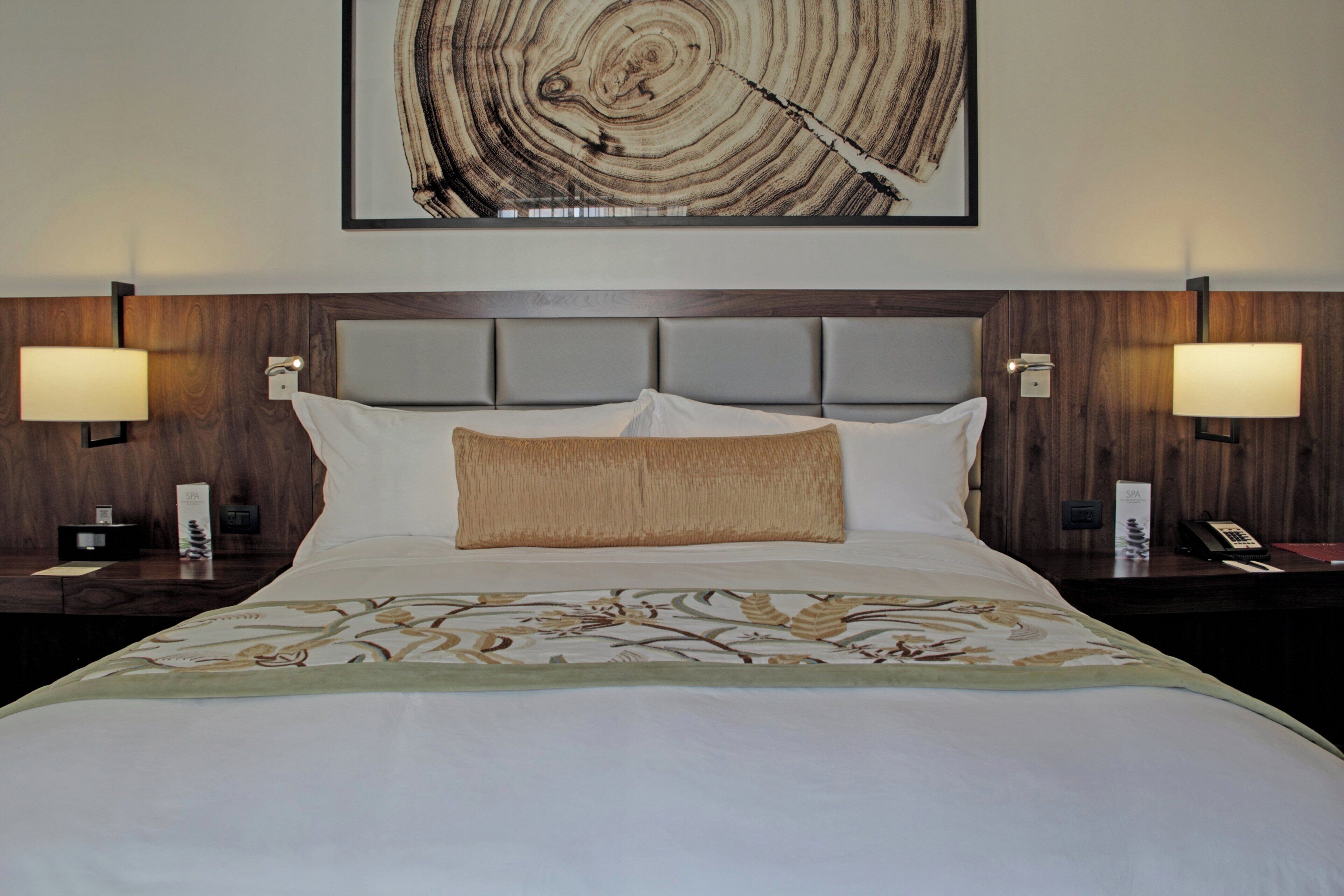 Habitación de hotel con una cama grande, dos lámparas de noche, un cabecero y una obra de arte colgada en la pared.