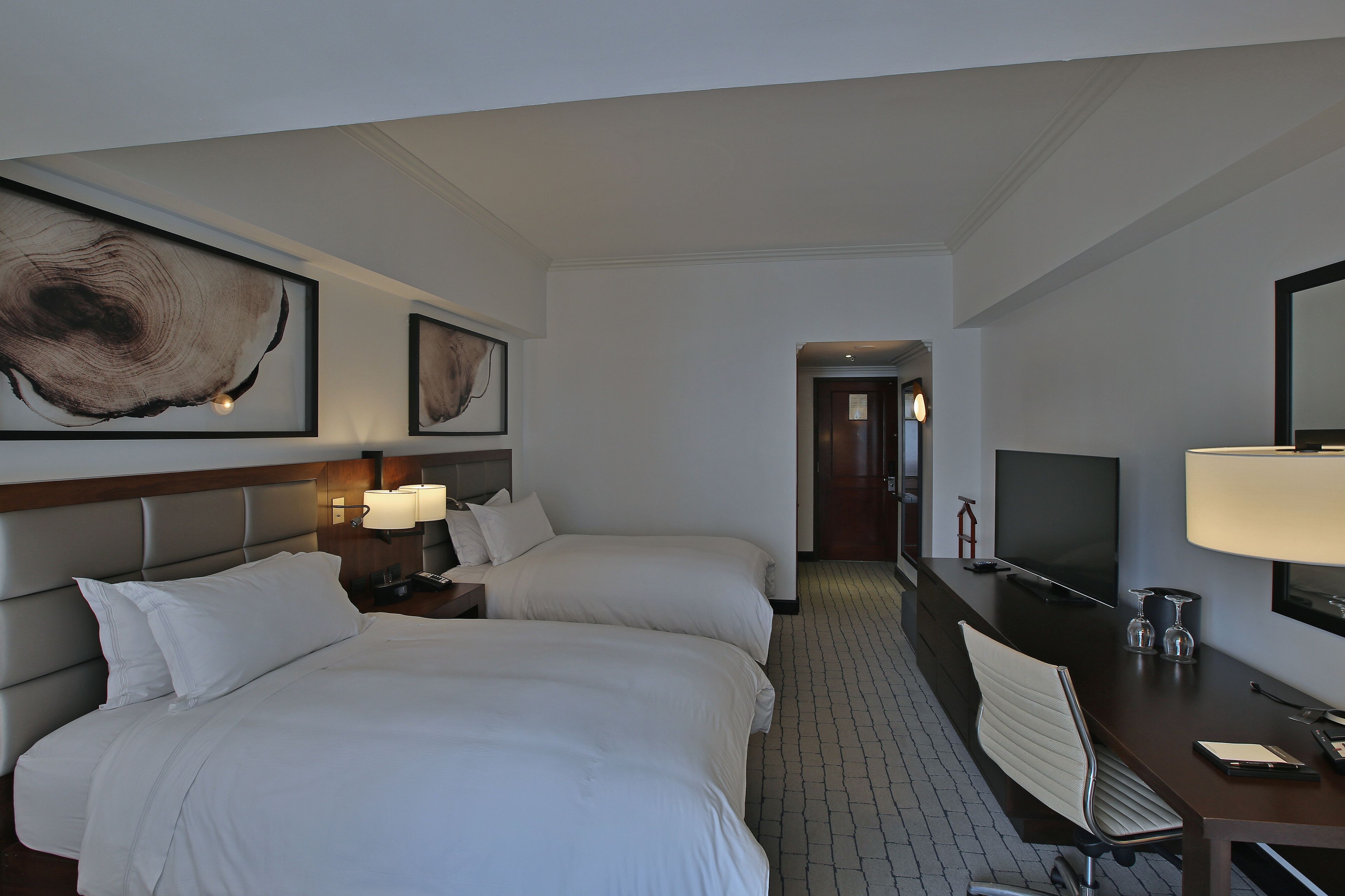 A hotel room with two beds, a desk with a TV, a lamp, and a large framed artwork on the wall.