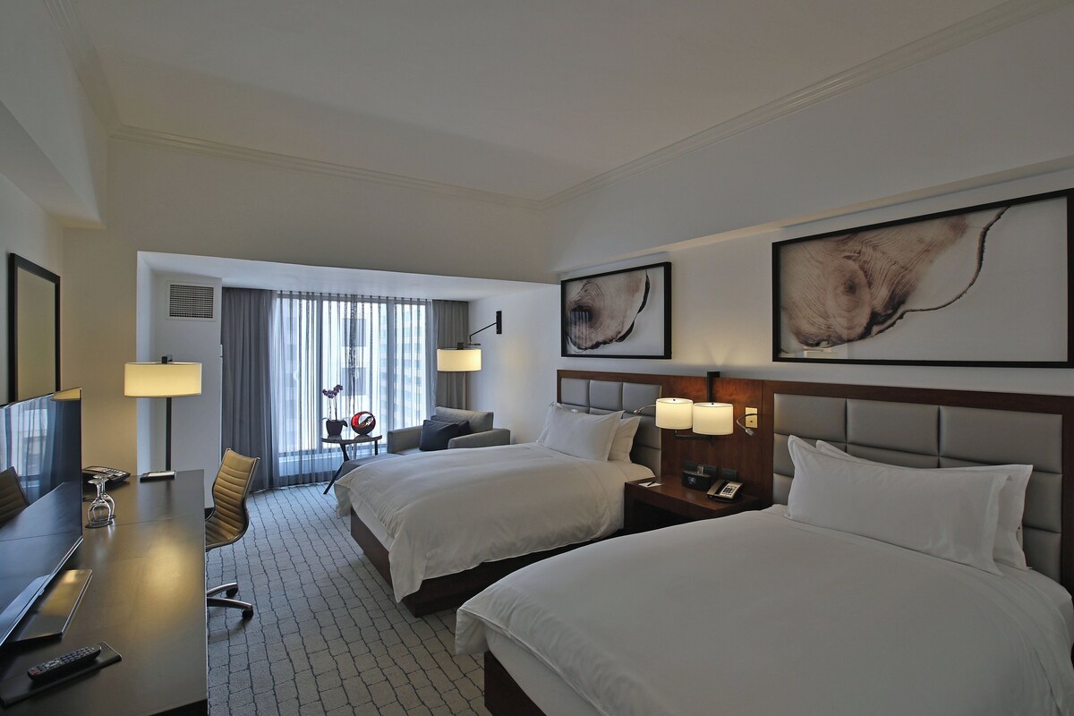 Habitación de hotel con dos camas, un escritorio con televisor, una silla y una ventana con cortinas.