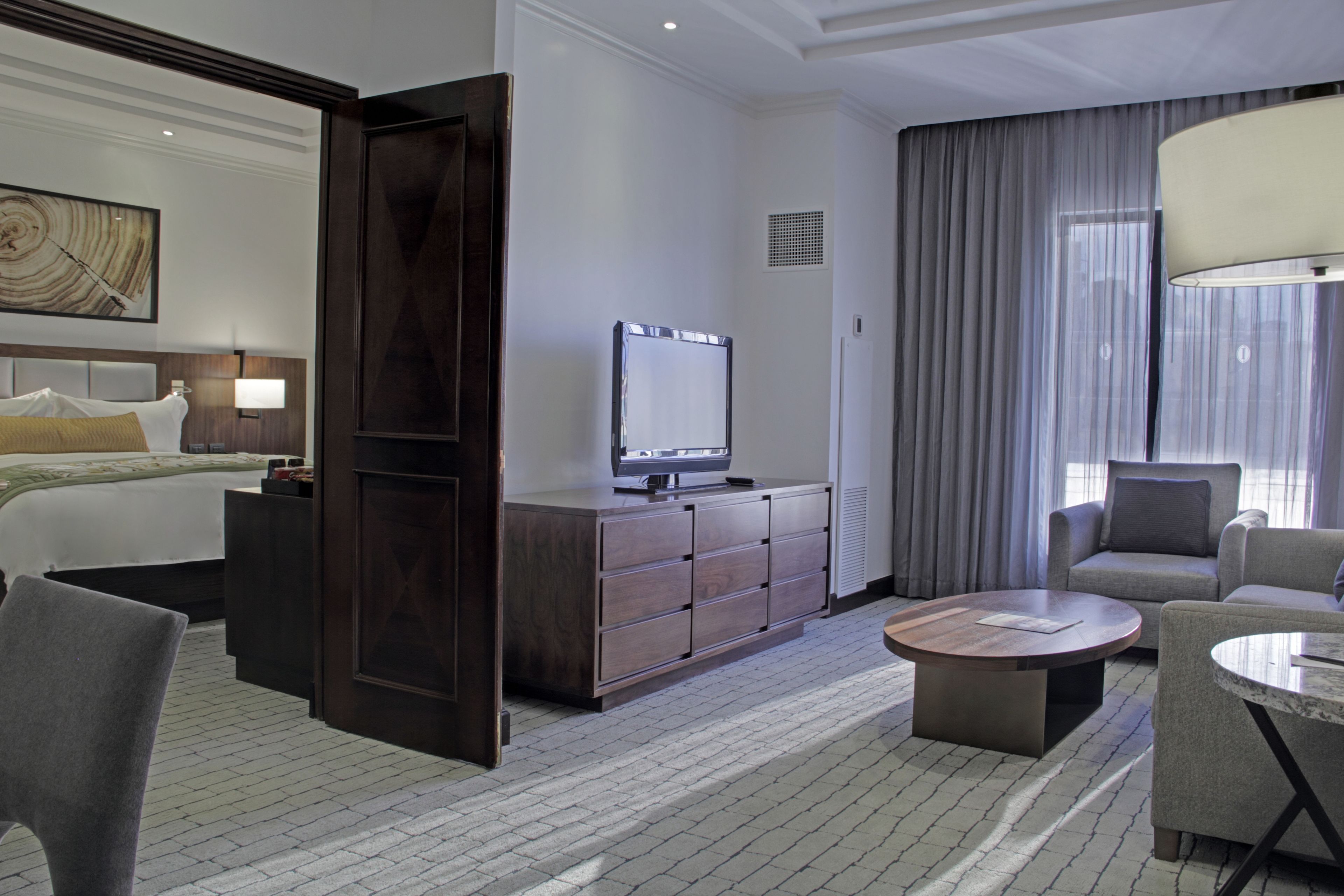 A hotel room with a bed, a TV on a wooden cabinet, a sofa, a coffee table, and a large window with curtains.