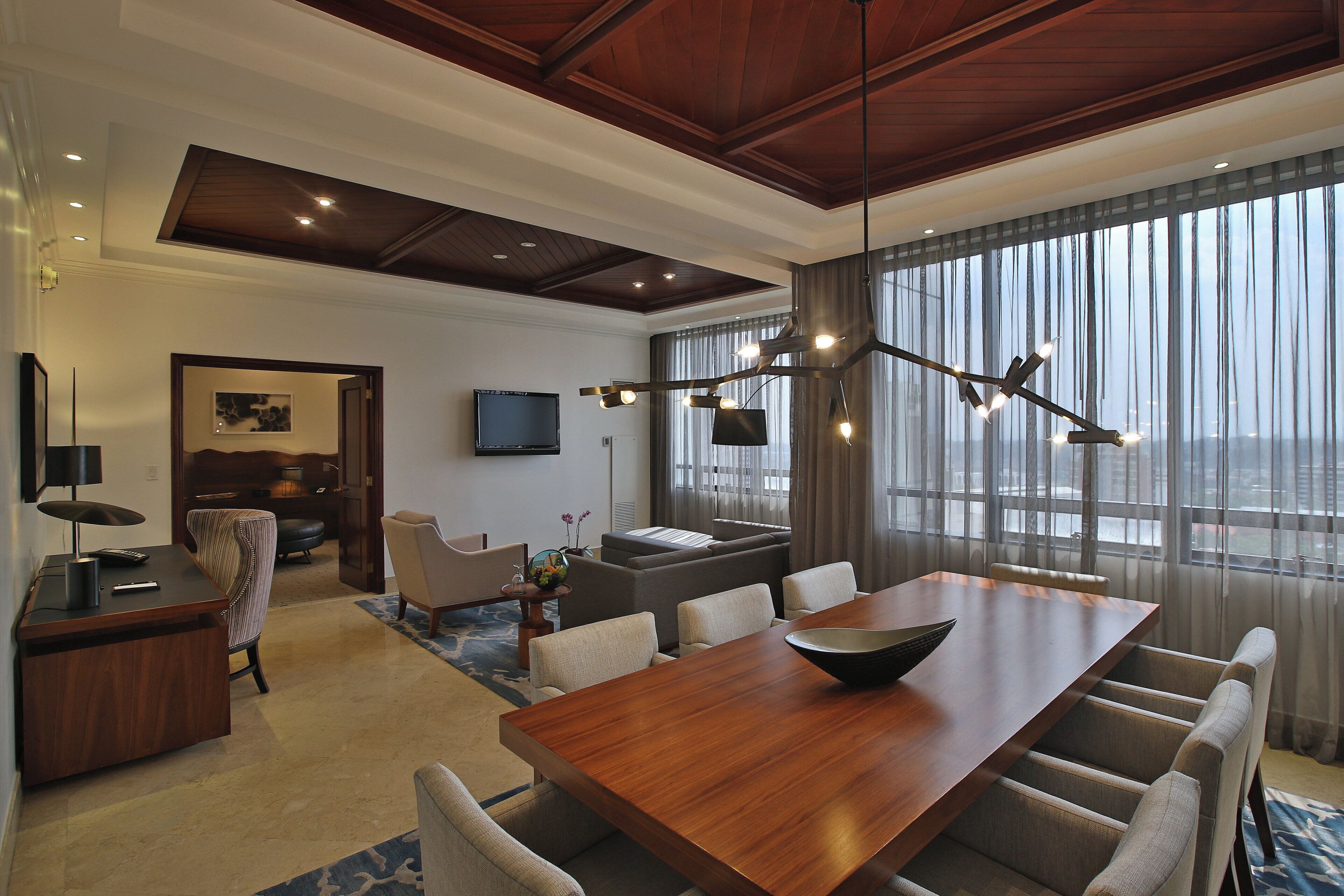 A modern hotel room with a wooden dining table, comfortable seating, a flat-screen TV, and a large window with curtains.