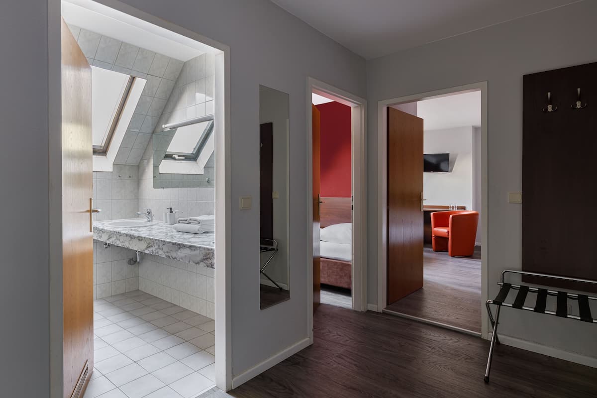 Une chambre d’hôtel moderne dotée d’une lucarne, d’une salle de bain avec un plan de toilette en marbre et d’un mur d’accent rouge.