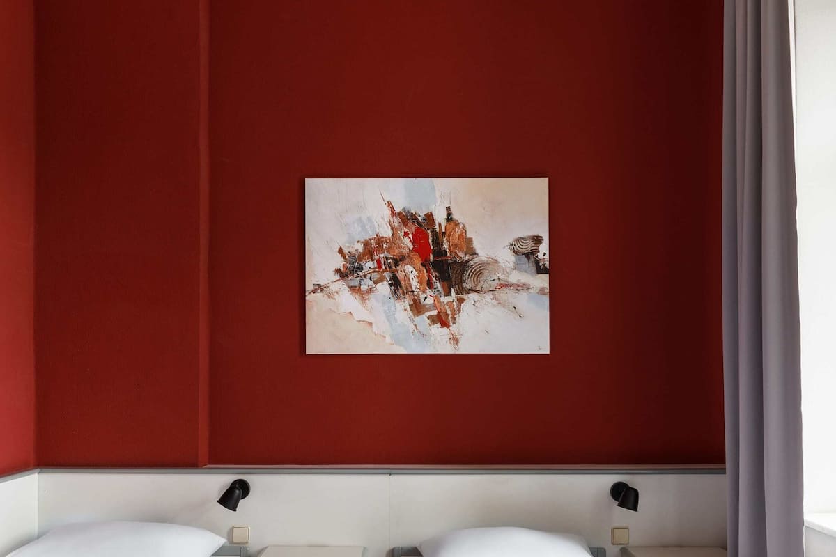 Une chambre d’hôtel avec deux lits simples, un mur d’accent rouge, un tableau et une fenêtre avec des rideaux.