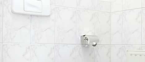 Casa de banho com sanita, um contentor de lixo, suporte para papel higiénico e uma planta num vaso.