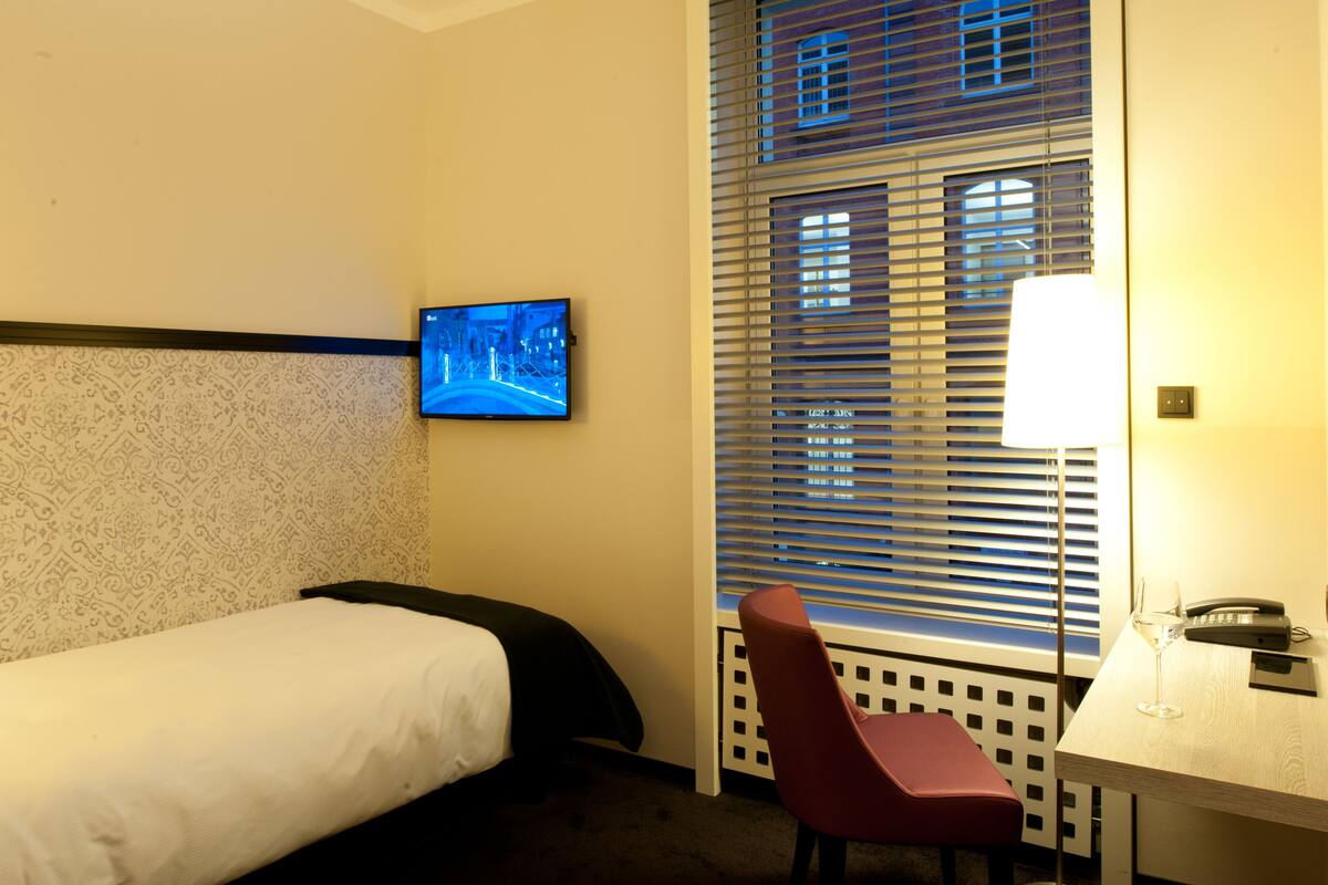 Una habitación de hotel con cama, escritorio con lámpara, silla y televisor en la pared.