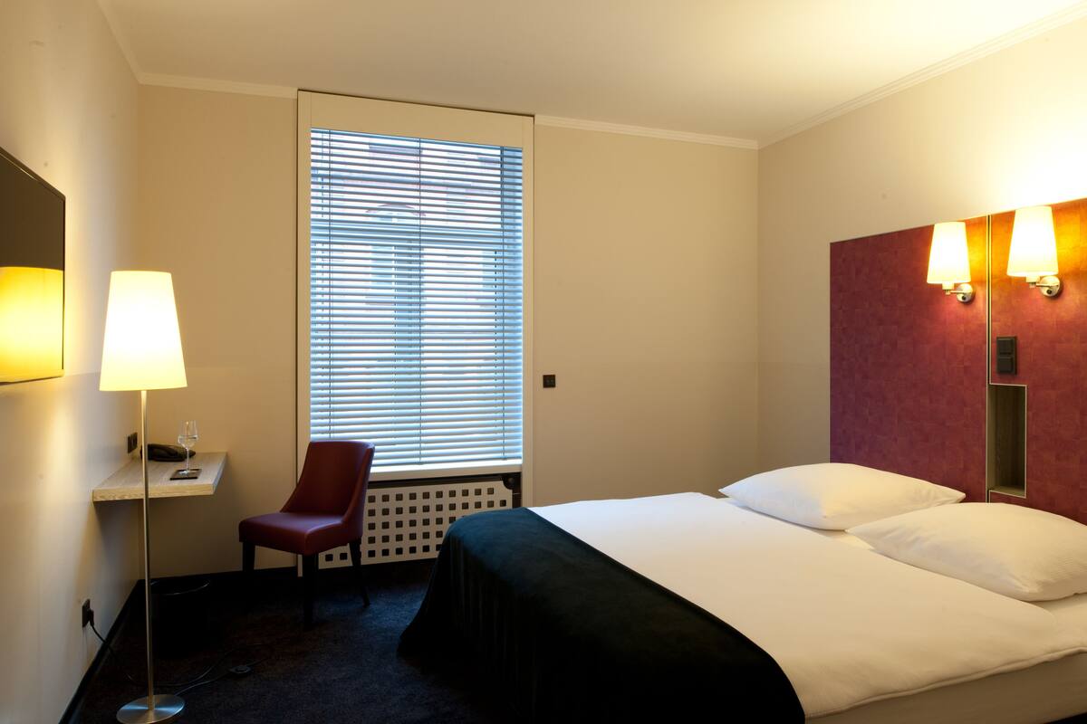 Una habitación de hotel con cama, una silla, un escritorio con lámpara y una ventana con persianas.