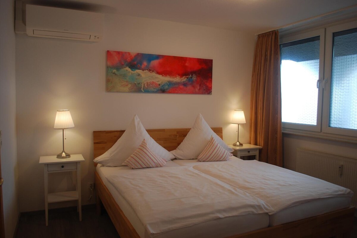 A hotel room with a bed, two bedside tables with lamps, a window with curtains, and a colorful abstract painting on the wall.