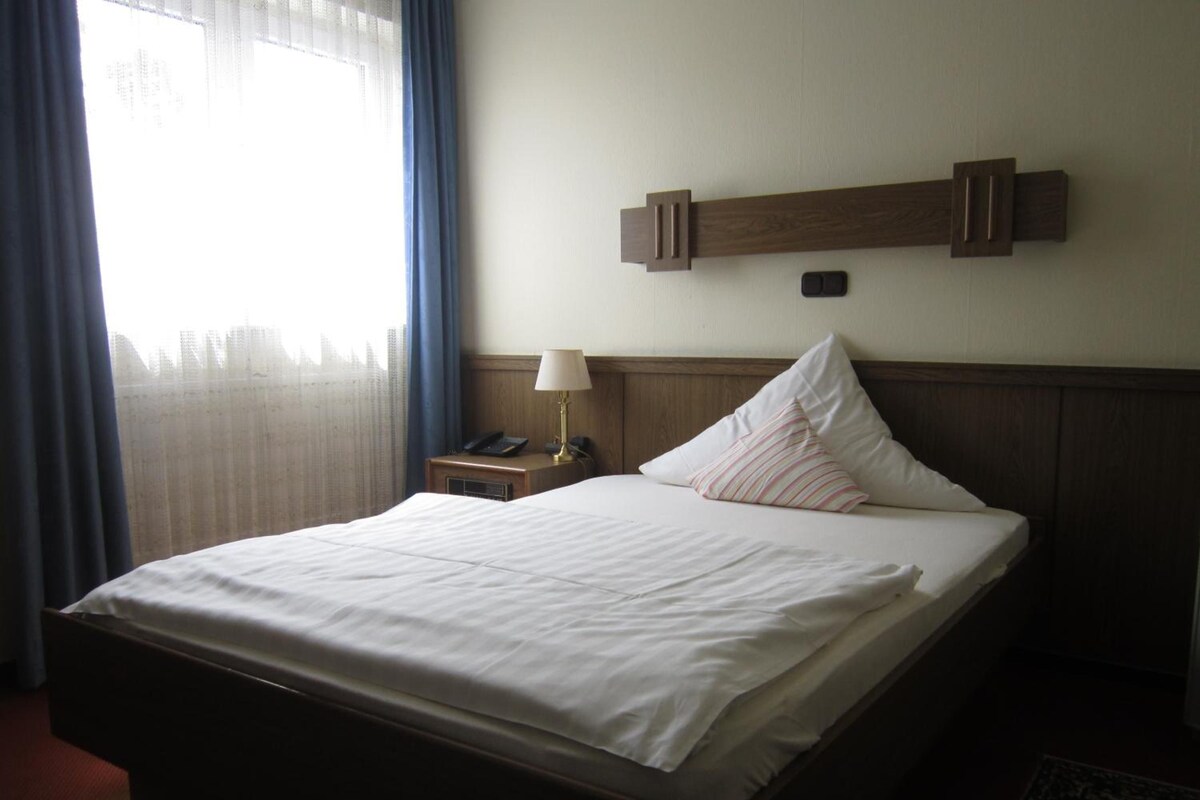 Ein Hotelzimmer mit einem Bett, einem Nachttisch, einer Lampe und einem Fenster mit Vorhängen.