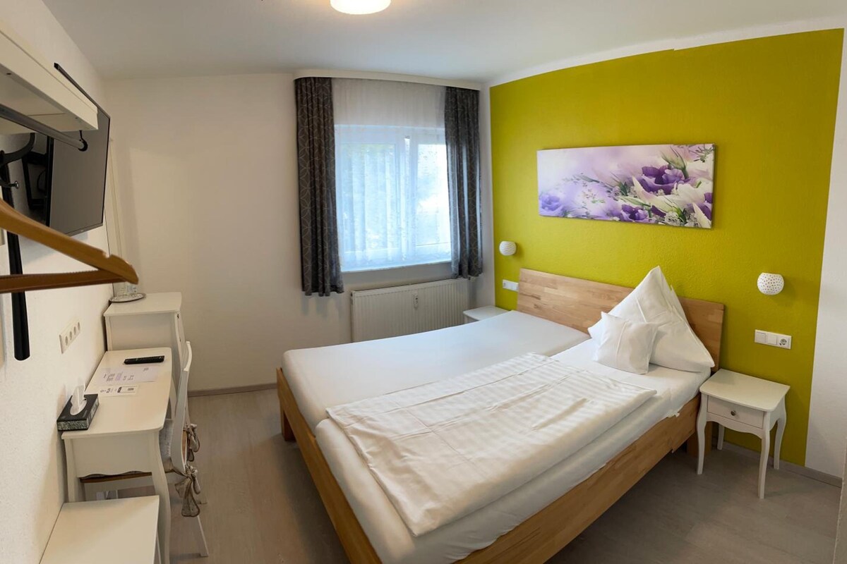A hotel room with a bed, a bedside table, a desk with a chair, a window with curtains, and a wall with a floral painting.