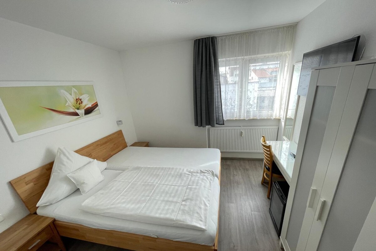 A hotel room with a single bed, a wooden nightstand, a wardrobe, a radiator, and a window with curtains.