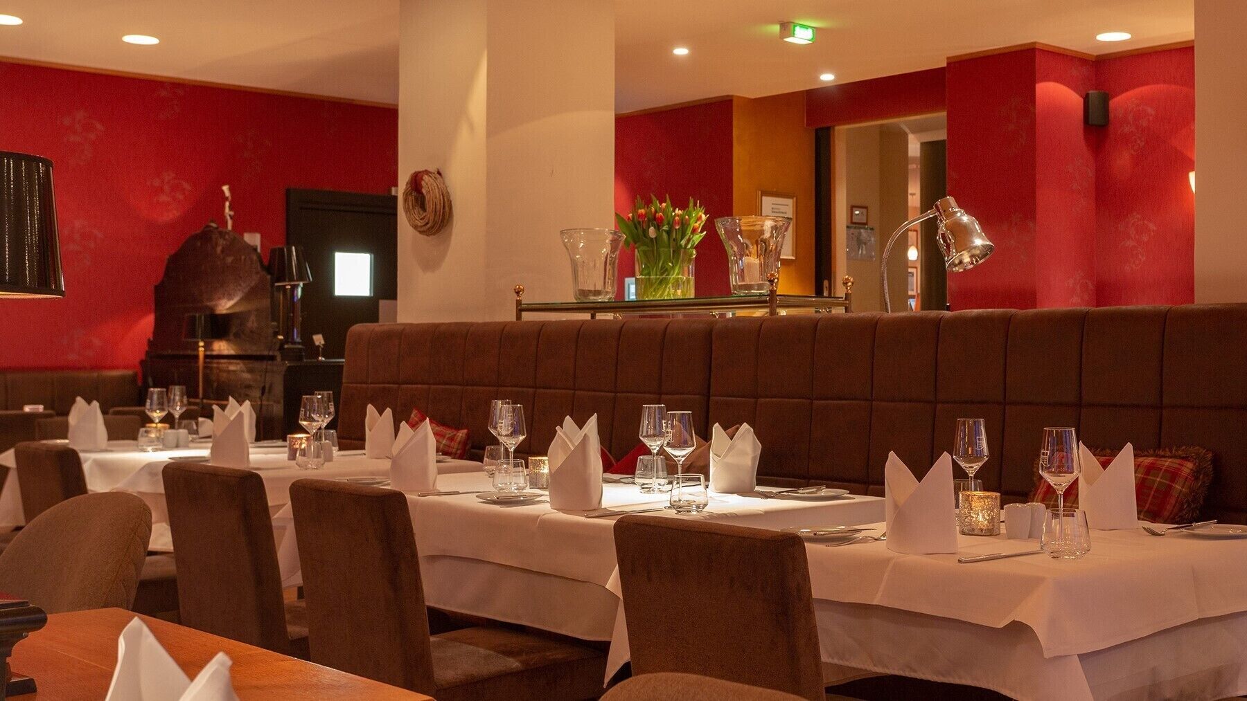 A restaurant with tables set for dining, featuring a red wall and a brown cushioned bench.