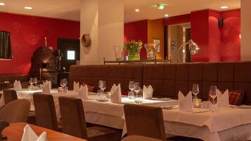 A restaurant with tables set for dining, featuring a red wall and a brown cushioned bench.