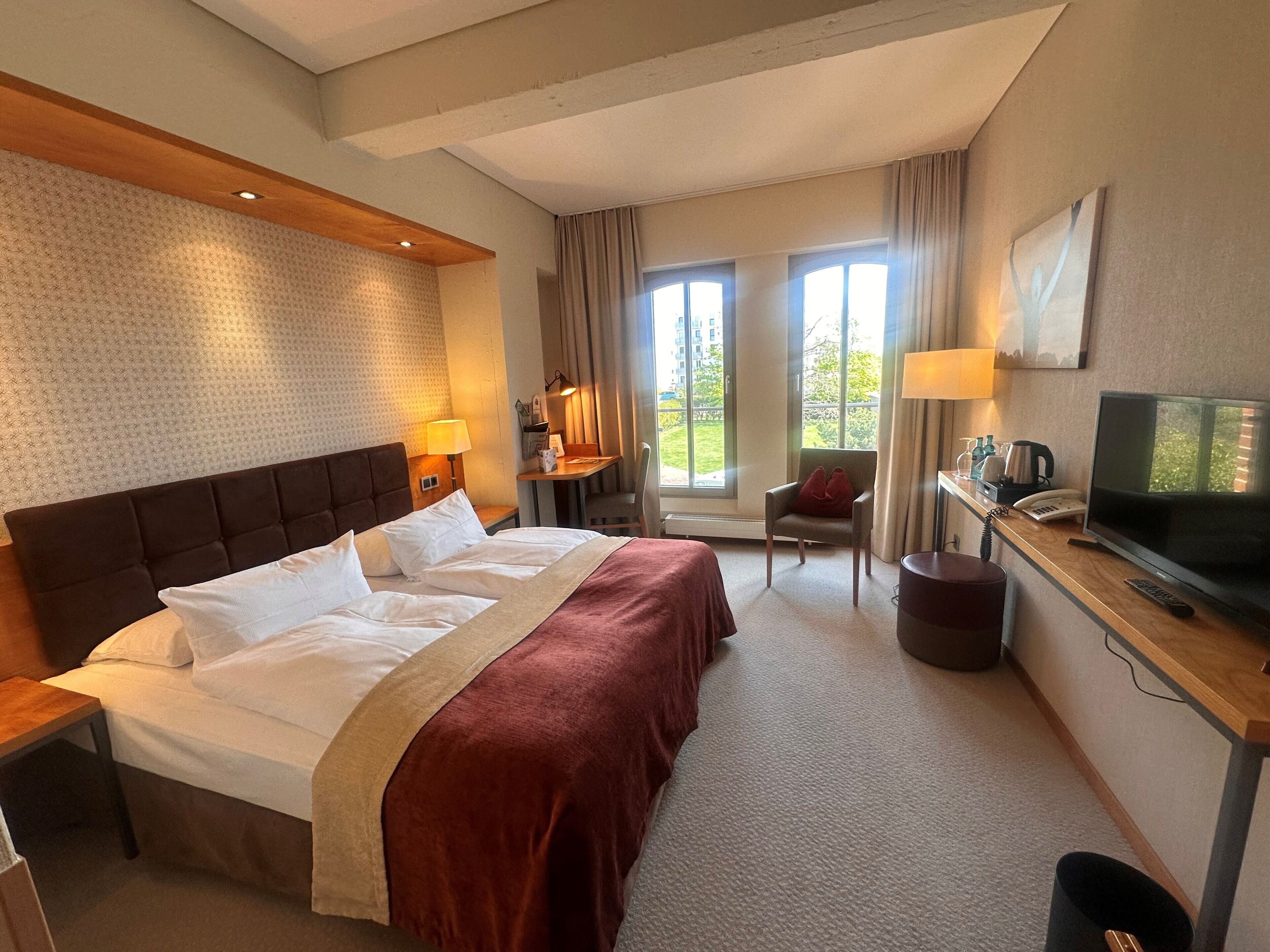 A hotel room with a large bed, a desk with a television, a chair, and a window with a view of the outdoors.