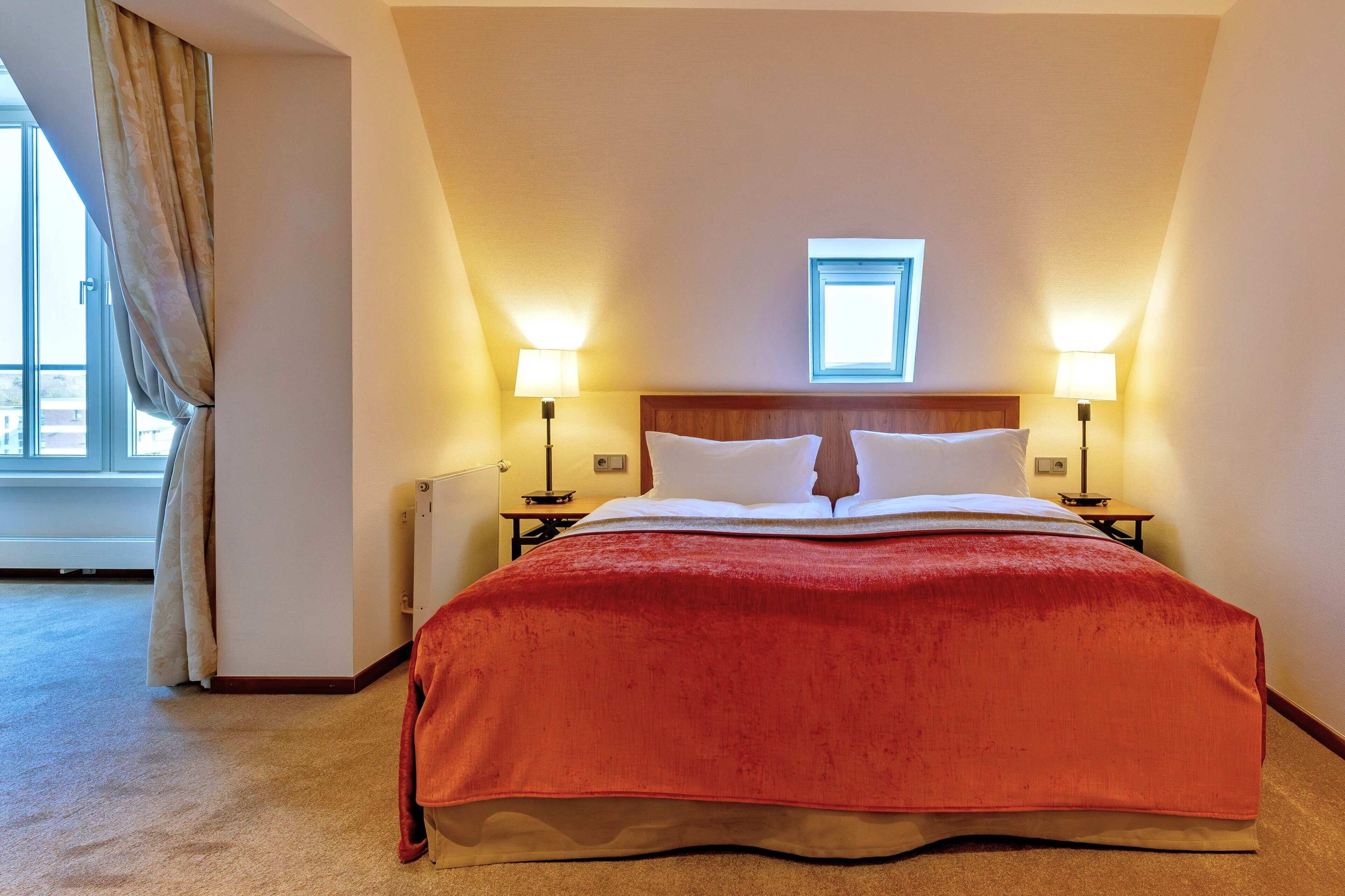 Una habitación de hotel con una cama grande, dos lámparas de noche, una ventana con cortinas y un tragaluz pequeño.
