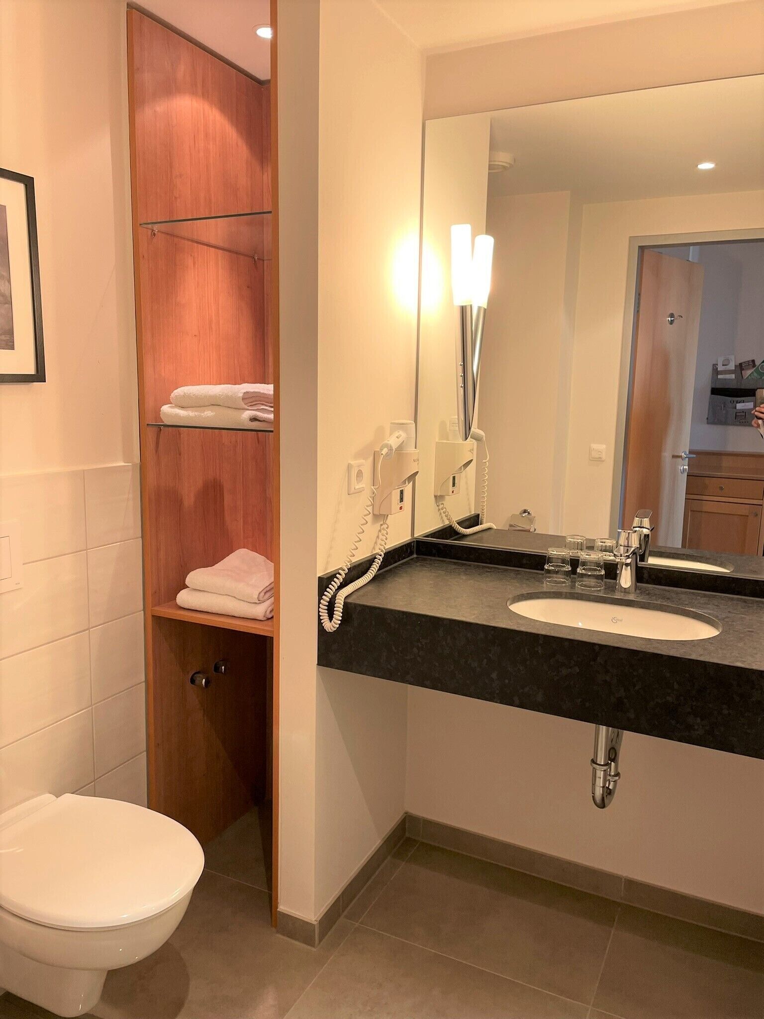 Une salle de bain moderne avec des toilettes blanches, un meuble-lavabo en bois doté d’un plan de travail noir, un grand miroir et un porte-serviettes.