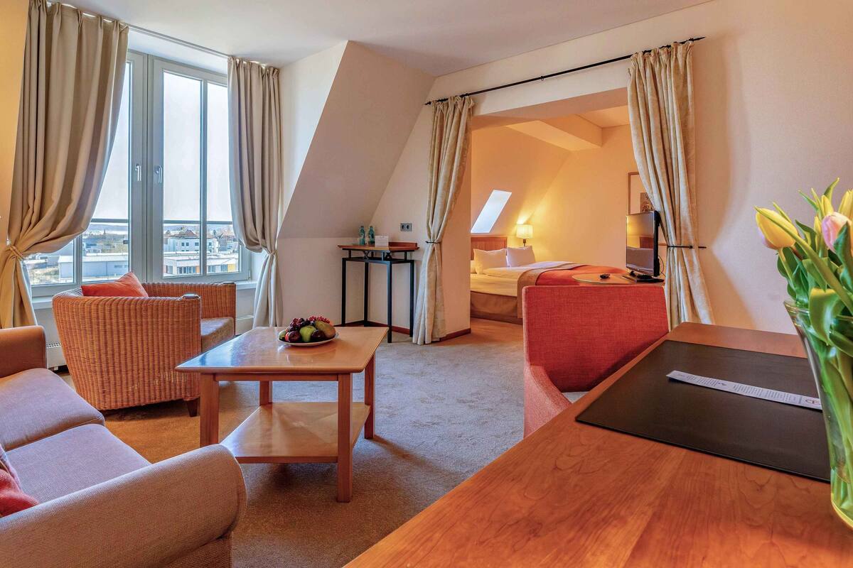 Een hotelkamer met een bank, een klein tafeltje met fruit, een rode stoel, een houten bureau en een uitzicht op de stad door grote ramen.