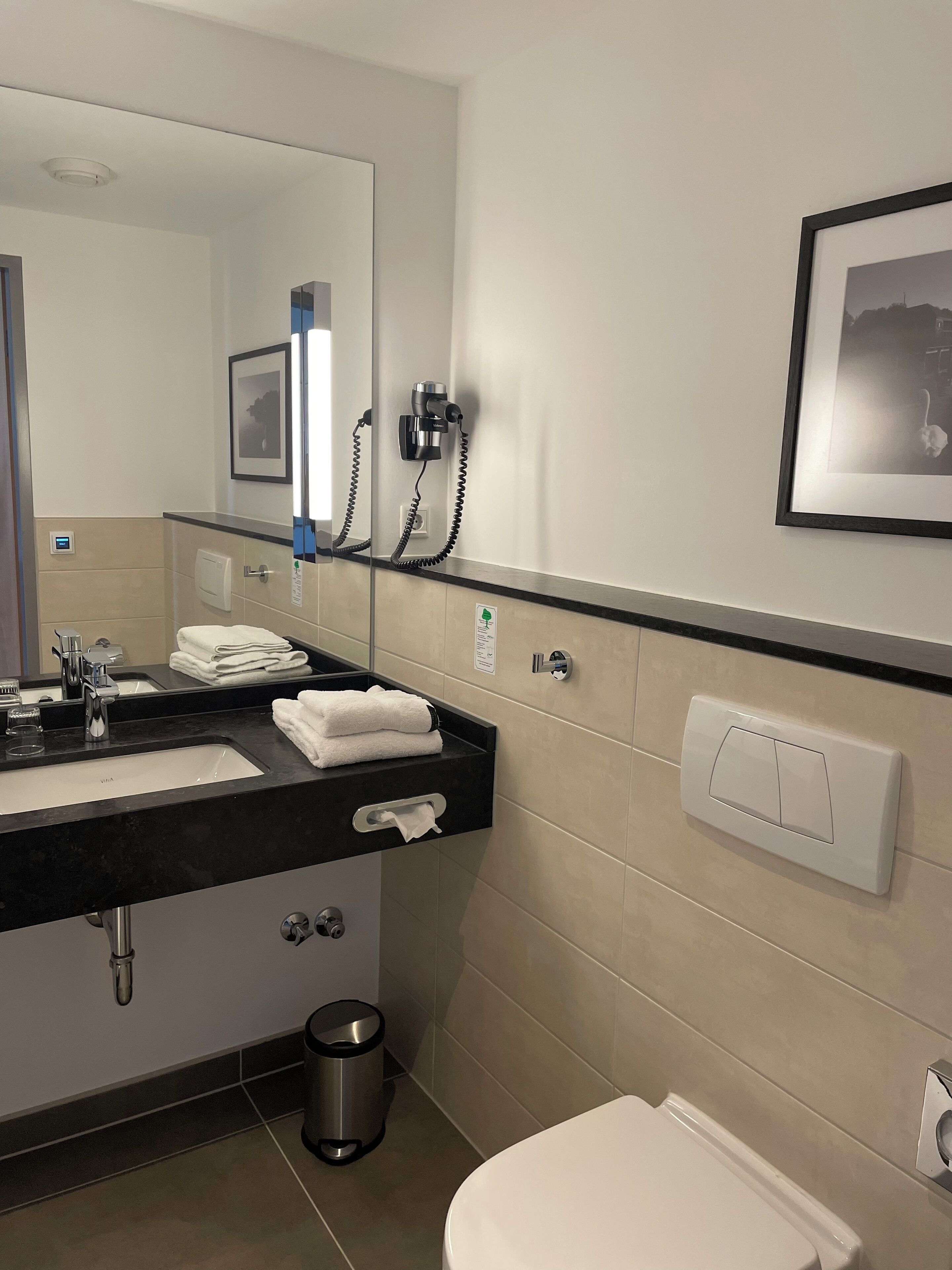 A modern bathroom with a black countertop sink, a mirror, a hair dryer, a trash can, and a toilet.