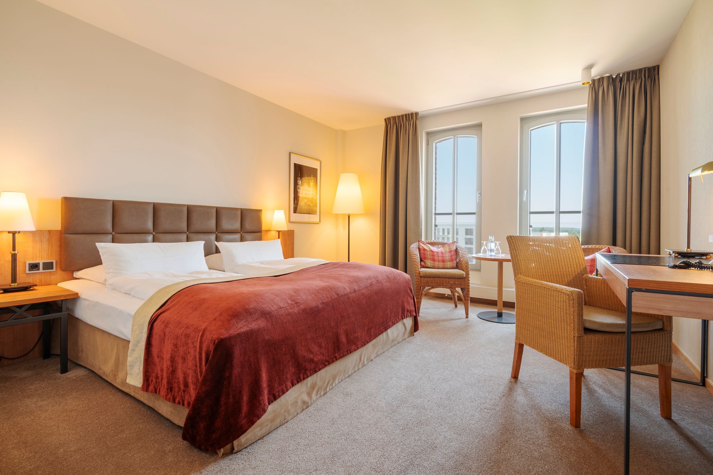 A hotel room with a large bed, two bedside lamps, a chair, a desk, and a window with curtains.