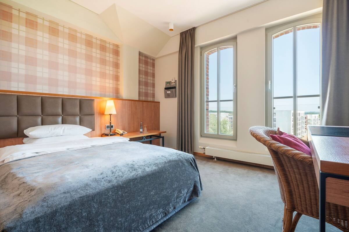 Een hotelkamer met een groot bed, een bureau met lamp, een rieten stoel en een raam met uitzicht.