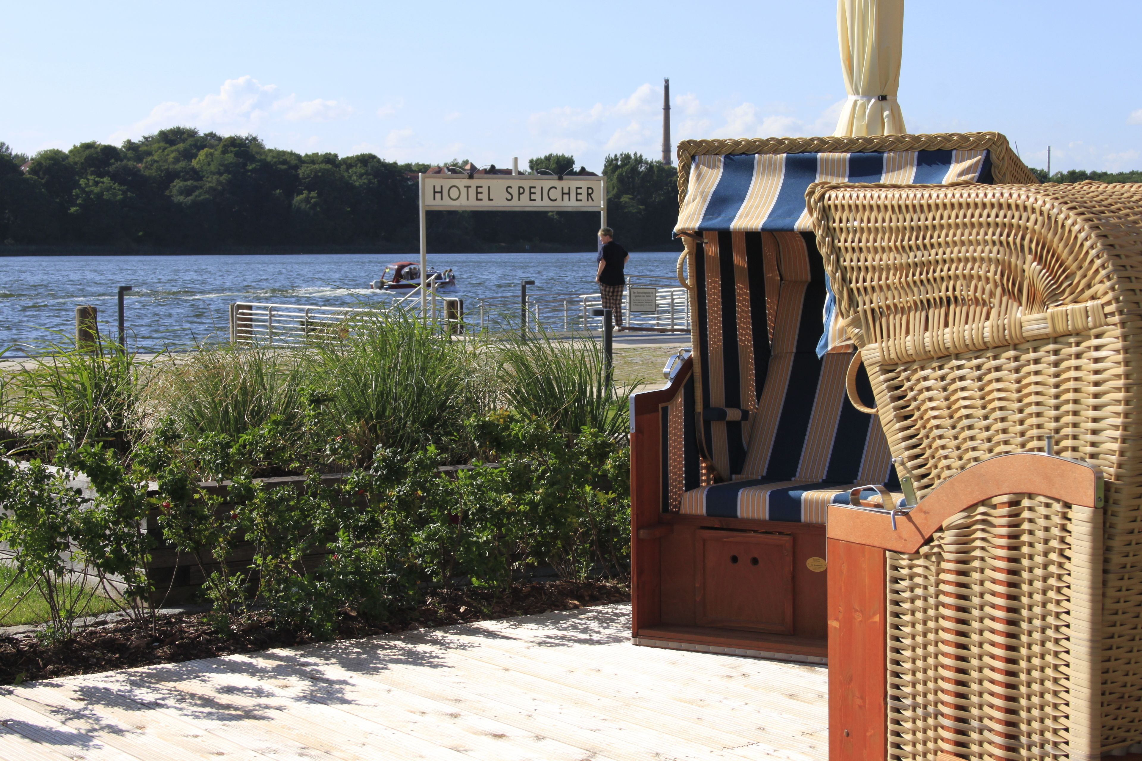 A wooden deck with a wicker chair and a sign reading "HOTEL SPEICHER" by the water.