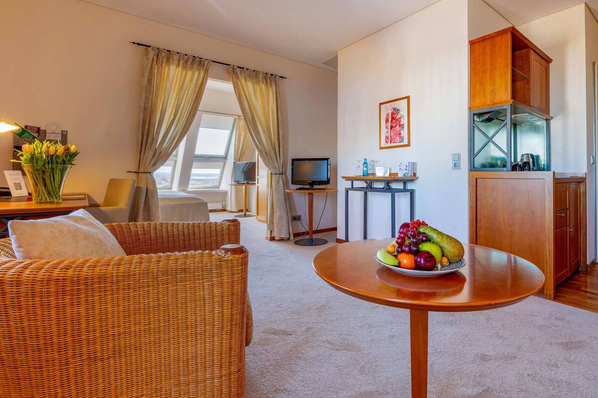 Hotelkamer met een rieten bank, een ronde tafel met fruit, een televisie, een klein keukenhoekje en een groot raam met gordijnen.