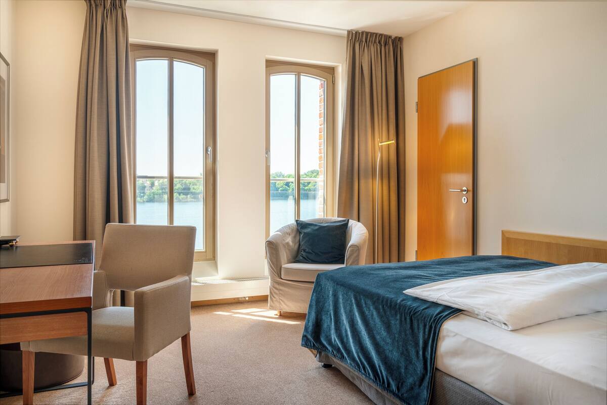 Een hotelkamer met een bed, een bureau, een stoel en uitzicht op een wateroppervlak door grote ramen.
