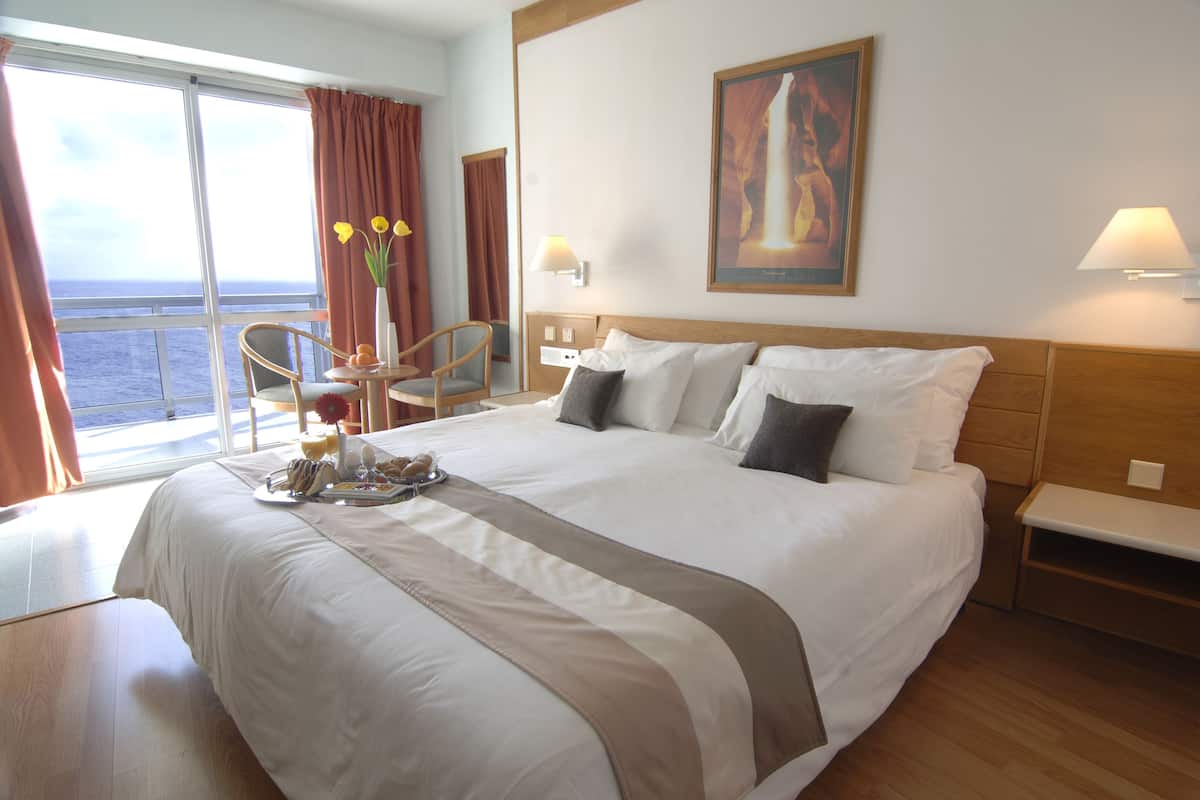 Habitación de hotel con una cama grande, una mesa de comedor con sillas, vista al mar y un cuadro en la pared.