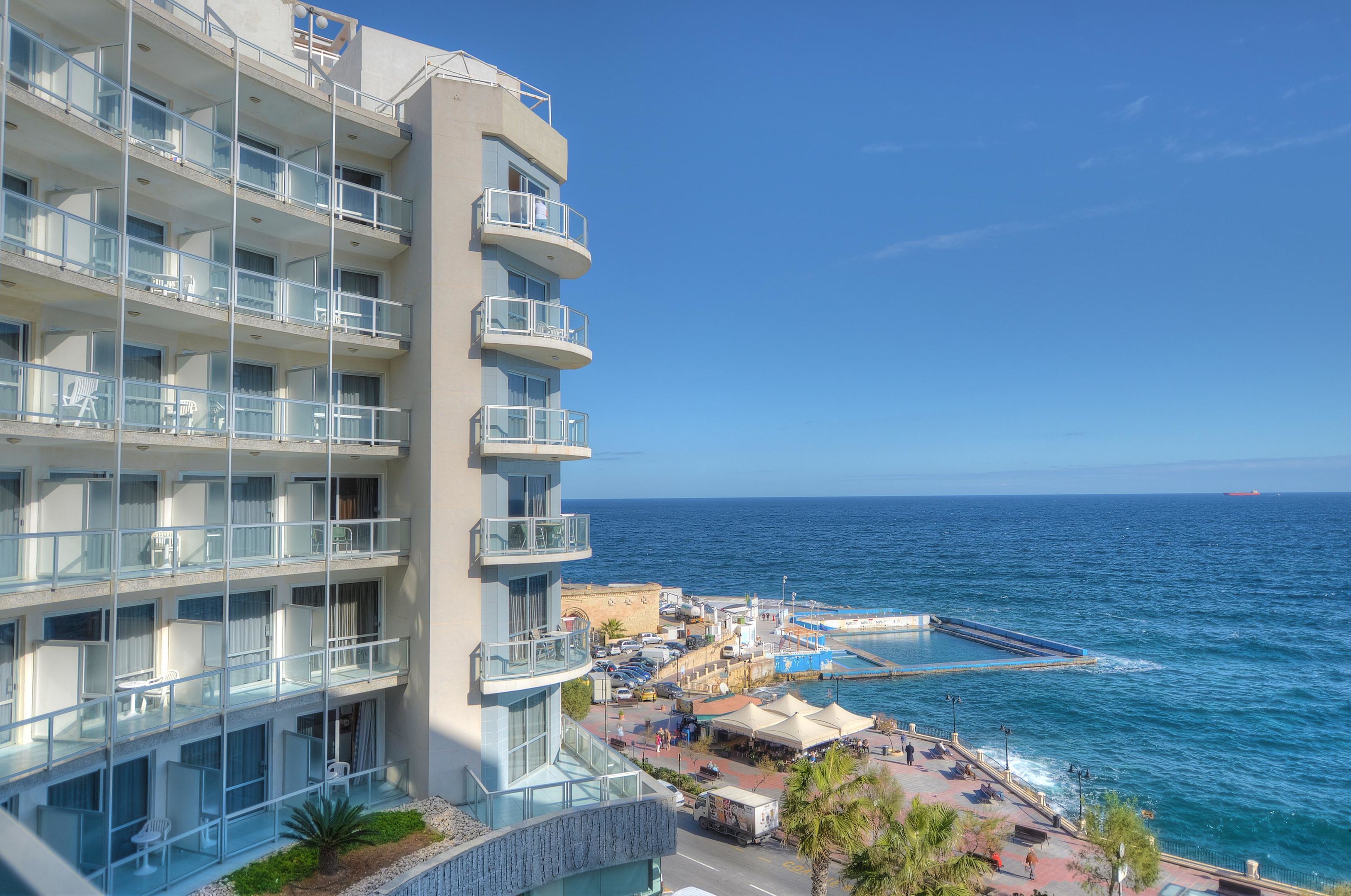 A multi-story hotel with balconies overlooking the sea, a dock area with boats, and a clear view of the ocean.
