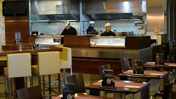 Ein Restaurant mit mehreren Tischen und Stühlen, einer Theke, an der zwei Köche arbeiten, und einer modernen Küche im Hintergrund.
