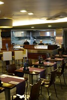 Ein Restaurant mit mehreren Tischen und Stühlen, einer Theke, an der zwei Köche arbeiten, und einer modernen Küche im Hintergrund.