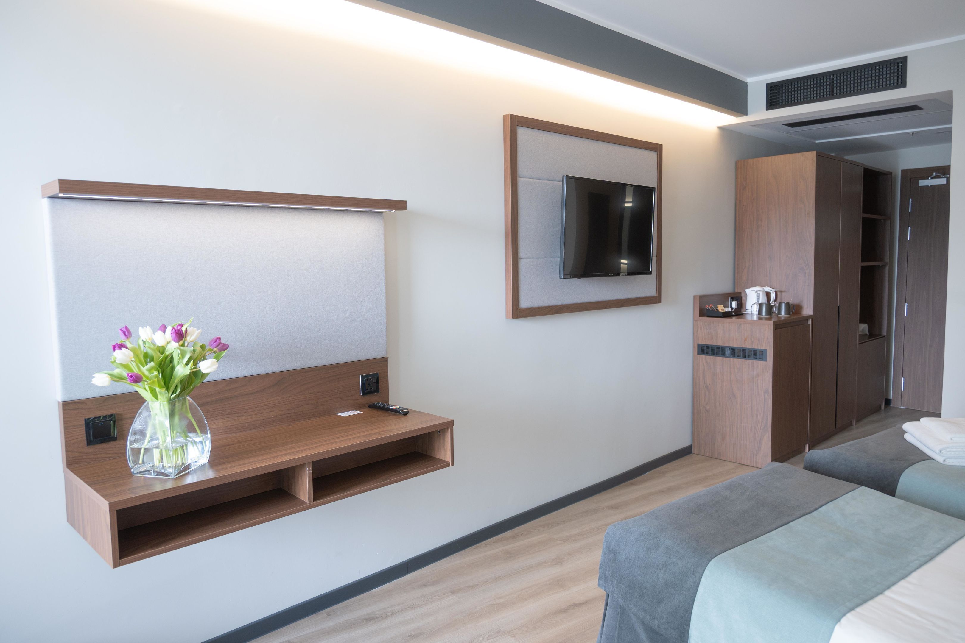 Quarto de hotel moderno com televisão de tela plana, um armário de madeira, uma mesa pequena com um vaso de flores e duas camas com roupa de cama cinza.