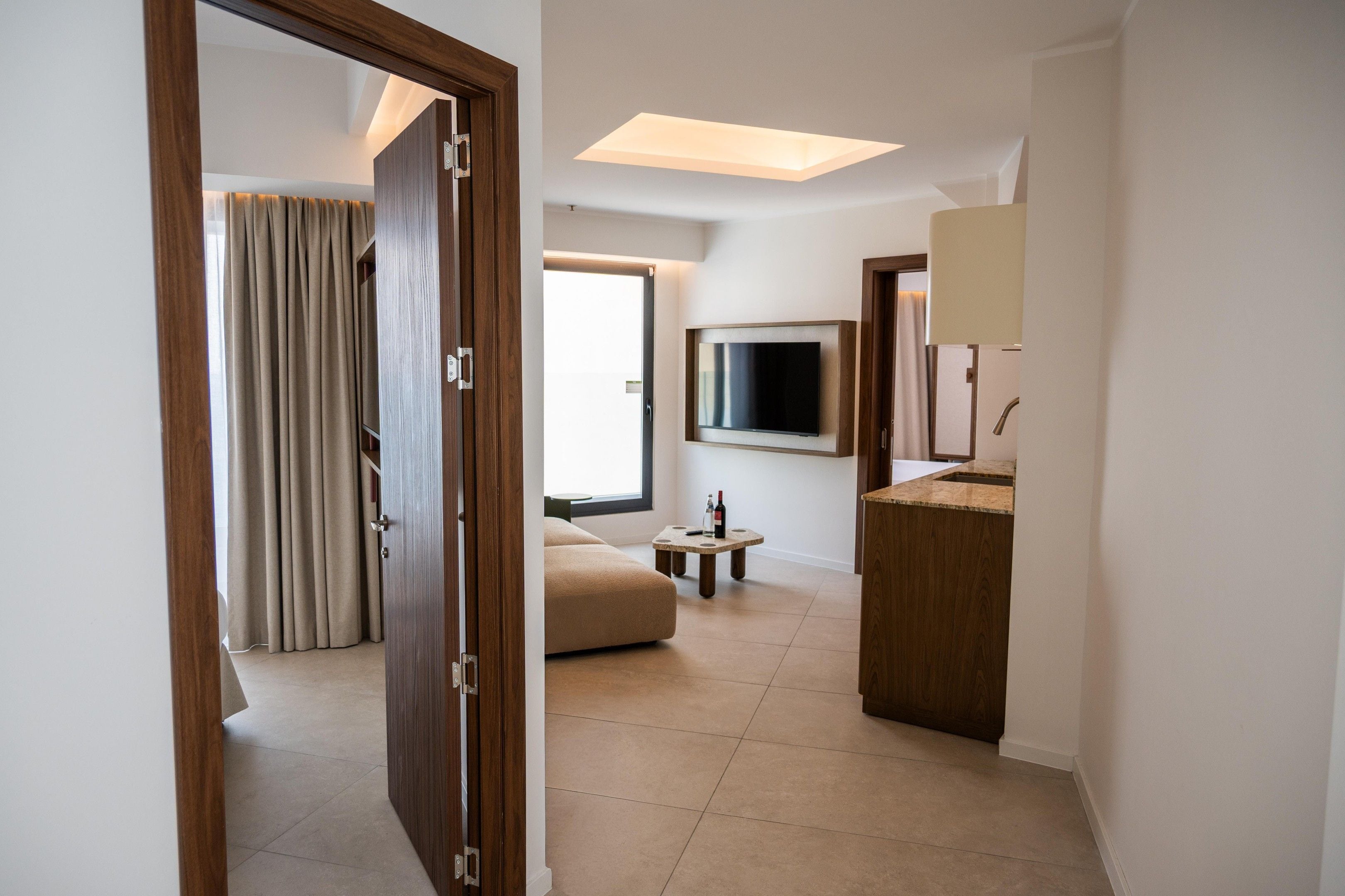 Una habitación de hotel con una puerta de madera, un televisor de pantalla plana montado en la pared, una mesita con una botella de vino y un sofá.