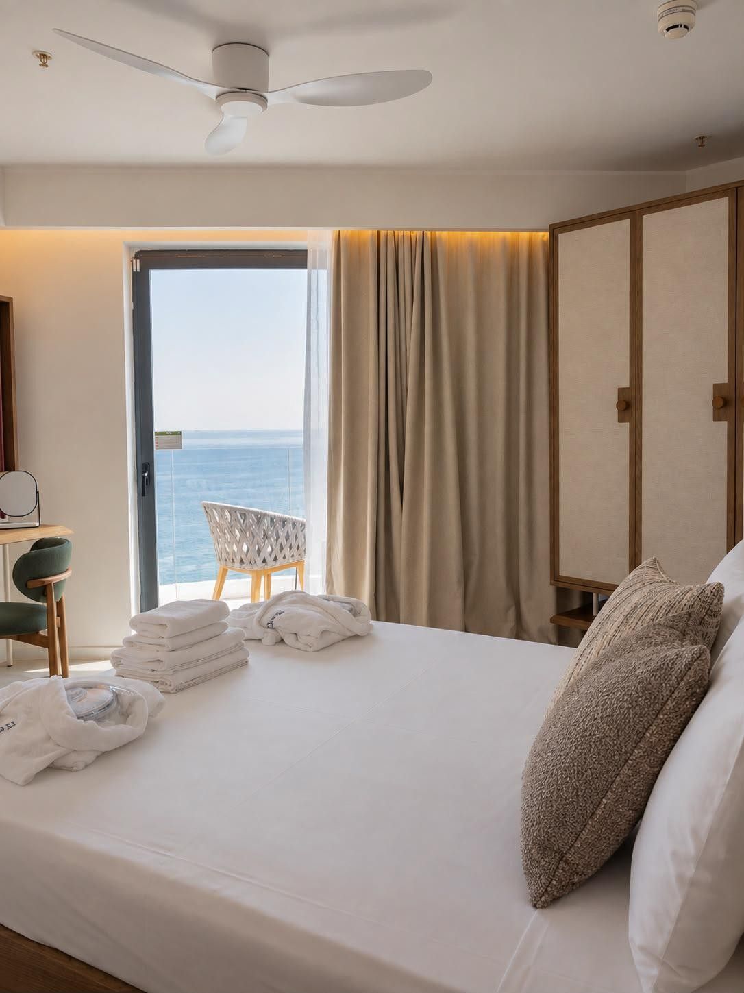 A hotel room with a bed, a chair, a table with towels, a view of the sea, and a ceiling fan.