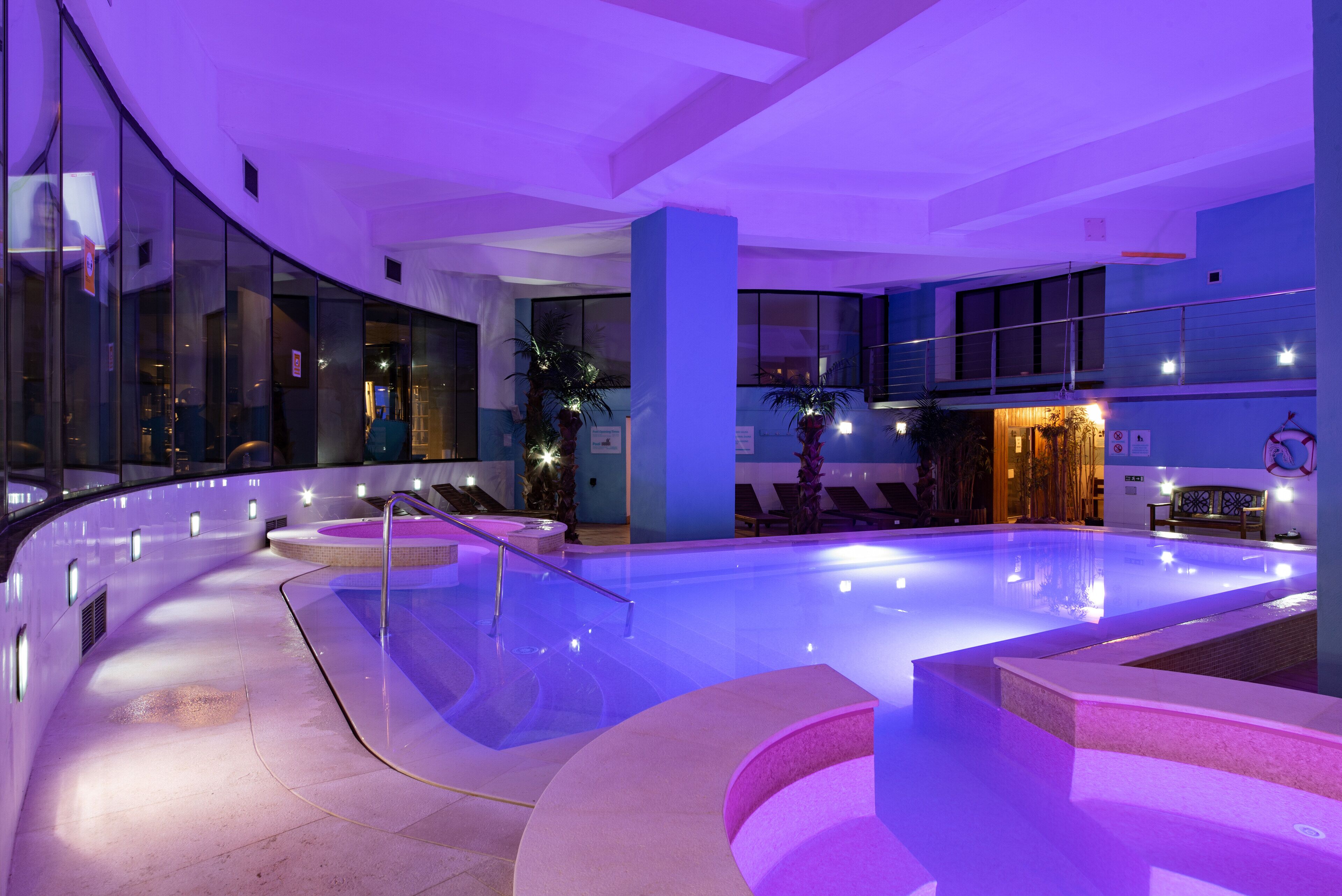 A modern indoor swimming pool with a curved design, glass walls, and ambient lighting.