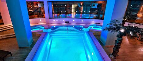 Una amplia piscina interior con iluminación azul, rodeada de paredes de vidrio y suelo de madera.