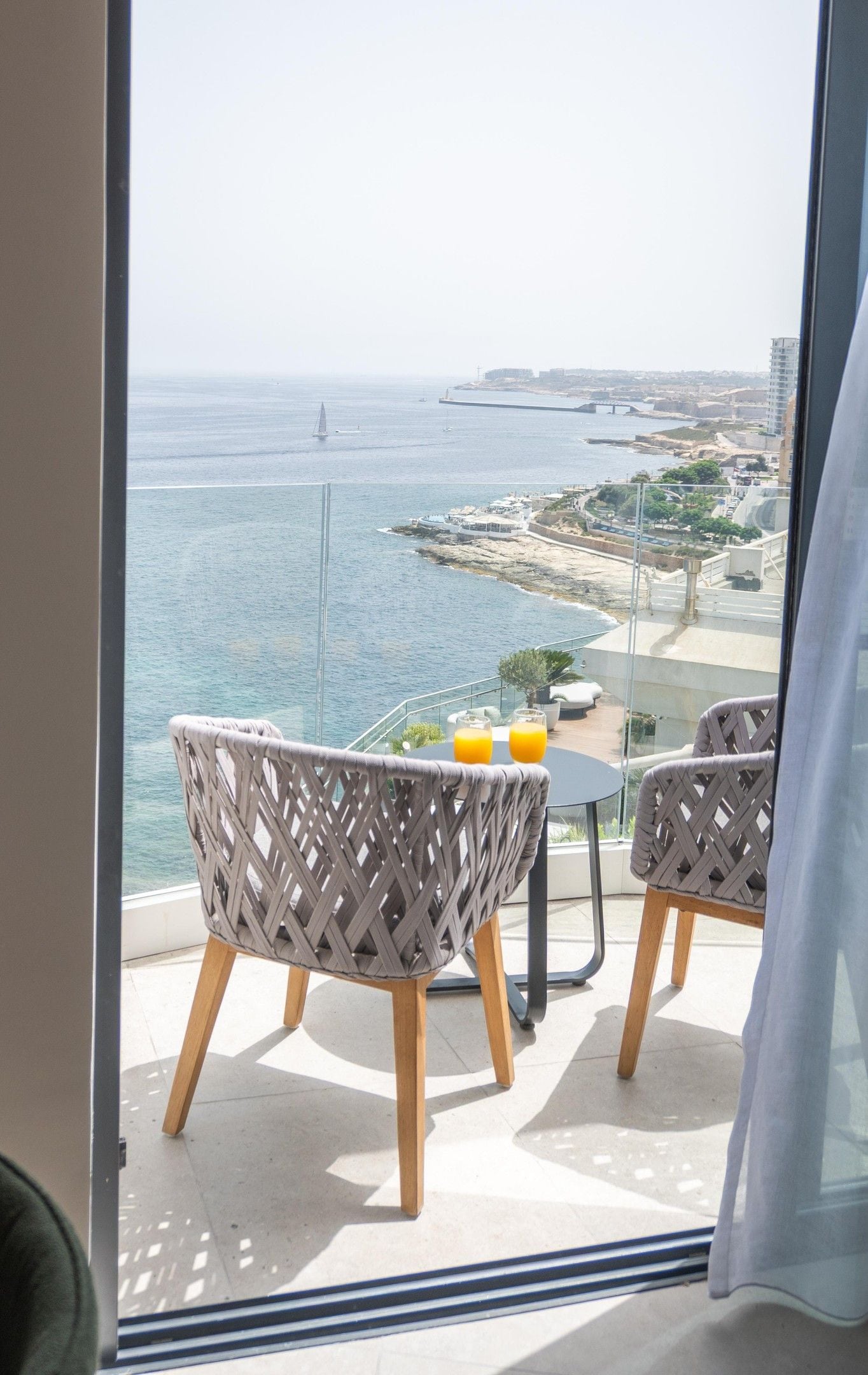 Un balcon avec deux fauteuils en osier et une petite table, d’où l’on peut admirer un paysage urbain côtier avec ses bâtiments et son port de plaisance.