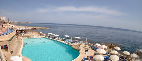 Un área de piscina con sillones de descanso y sombrillas, una piscina azul cristalina y vistas al mar.