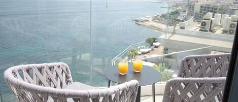 A balcony with wicker chairs, a small table with two glasses of orange juice, and a view of the sea and cityscape.