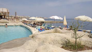 A pool area with lounge chairs and umbrellas, surrounded by large rocks and a view of the sea.