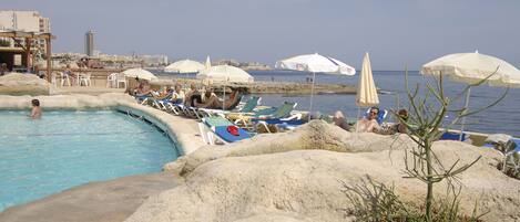 A pool area with lounge chairs and umbrellas, surrounded by large rocks and a view of the sea.