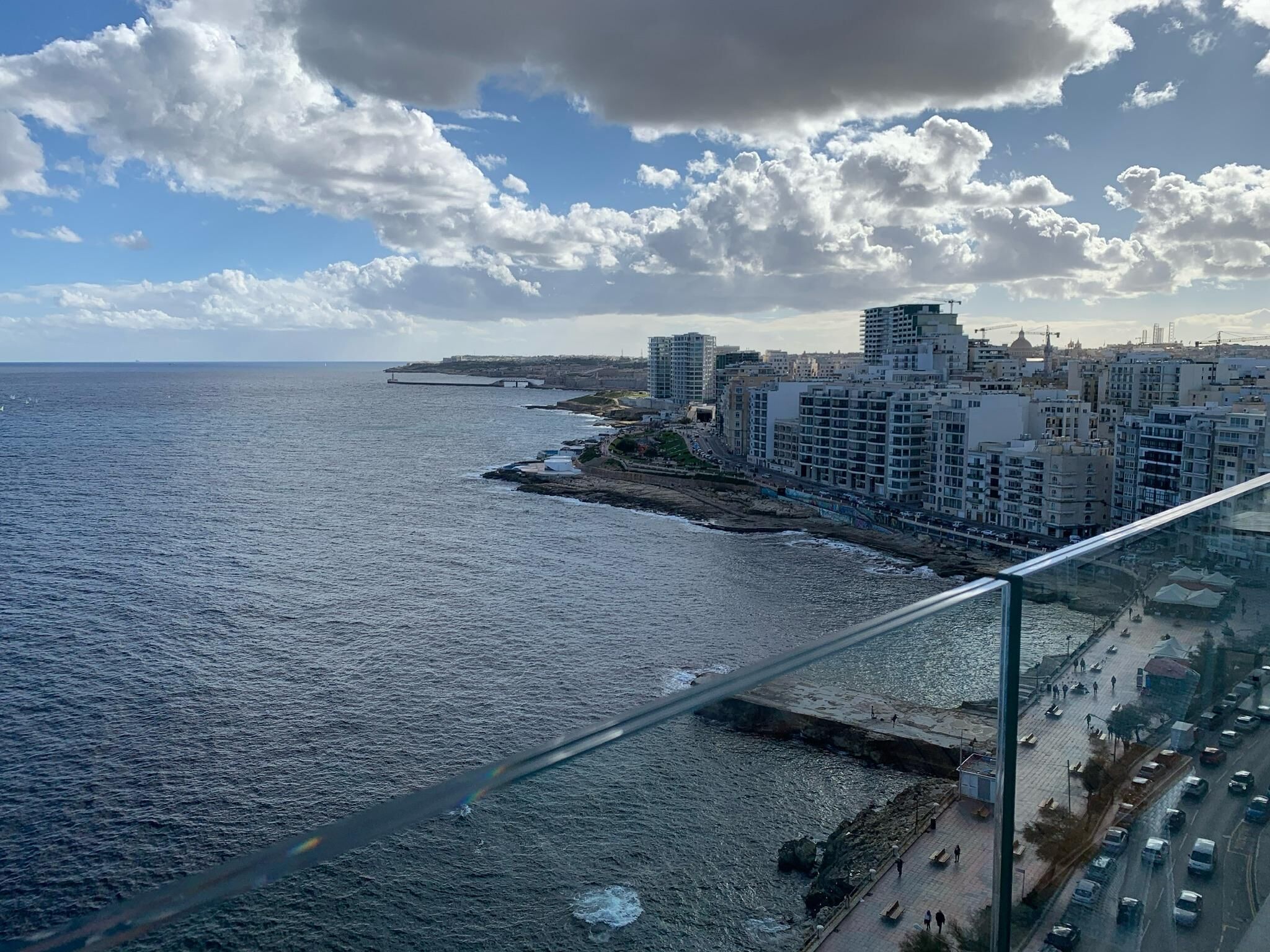 A coastal cityscape with high-rise buildings, a promenade, and a view of the sea.