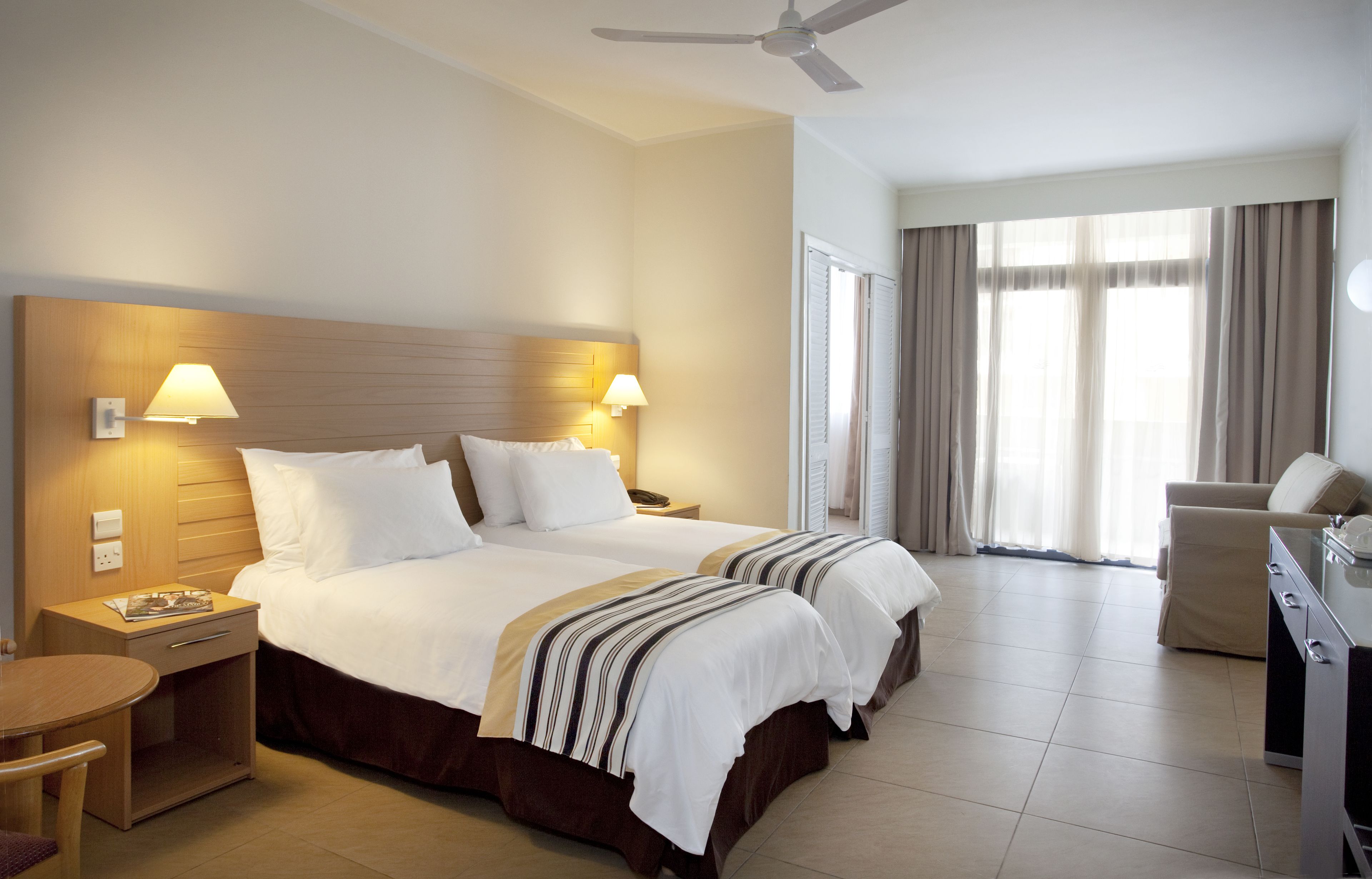 A hotel room with two beds, a desk, a chair, a ceiling fan, and large windows with curtains.