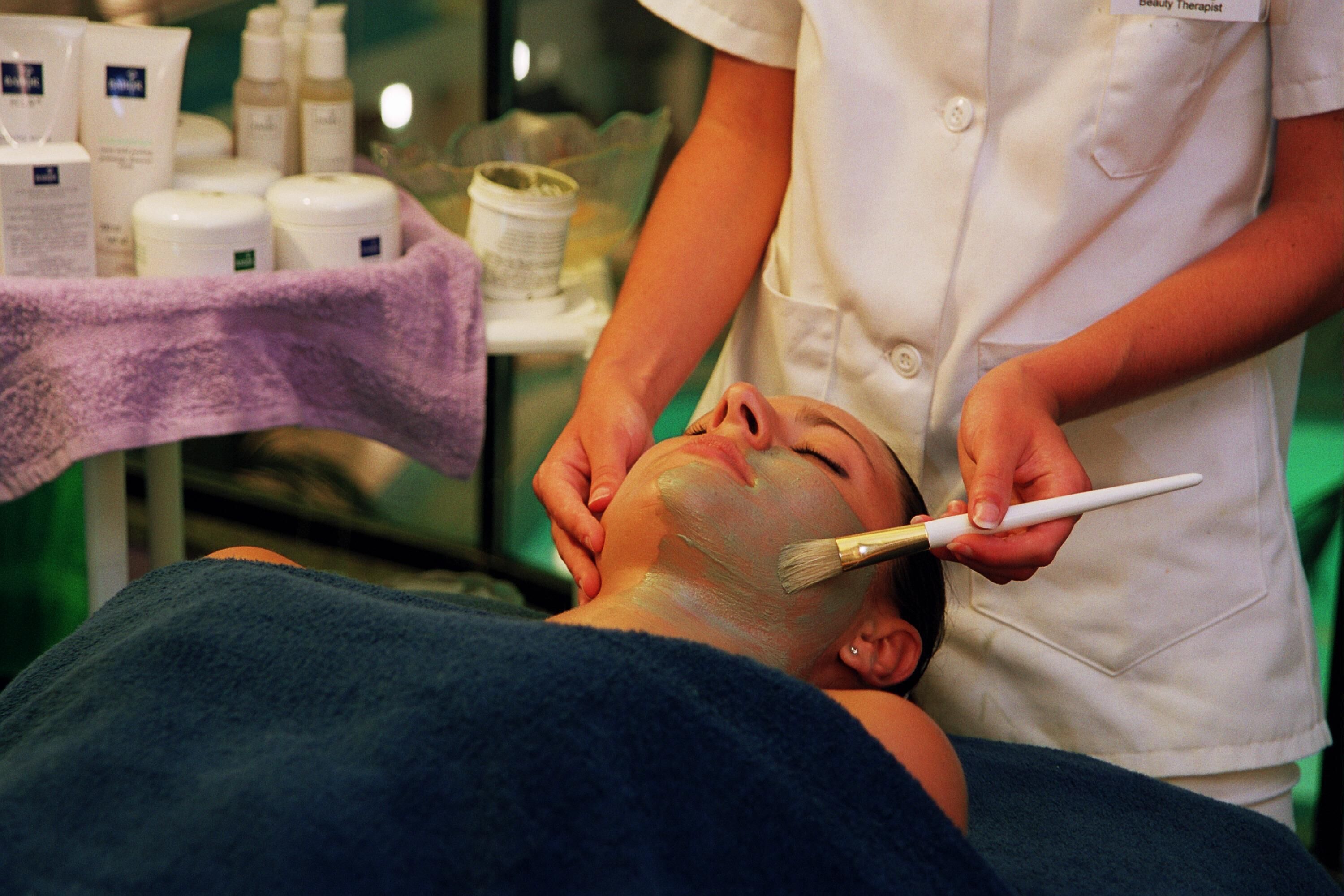 A person receiving a facial treatment with a brush, surrounded by various skincare products.