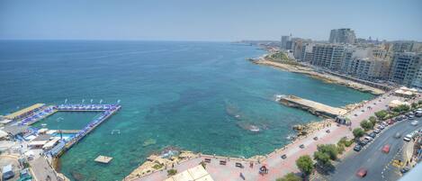 A coastal cityscape with a clear view of the sea, a pier, and a road with parked cars.