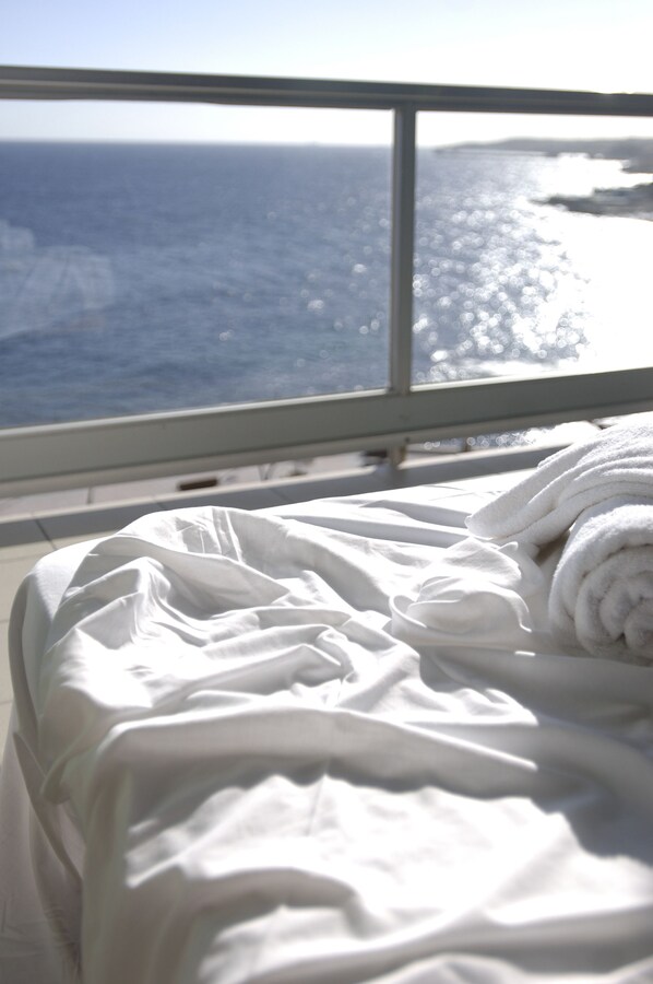 Una cama con sábanas blancas y vista al océano a través de una ventana.