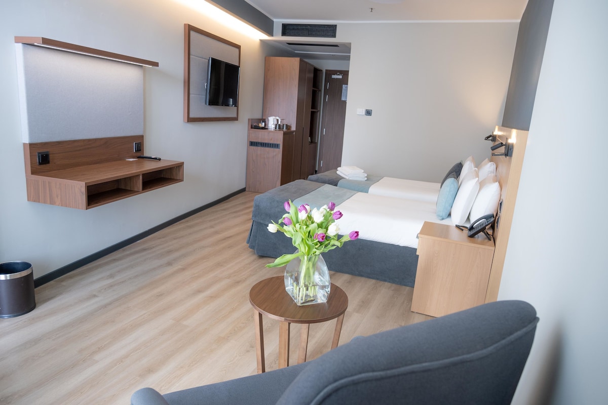 A modern hotel room with a bed, bedside table, and a small table with a vase of flowers.
