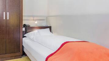 Una cama individual con ropa de cama blanca y una manta naranja, un armario de madera, un teléfono en la mesita de noche y un portalámparas fijado a la pared.