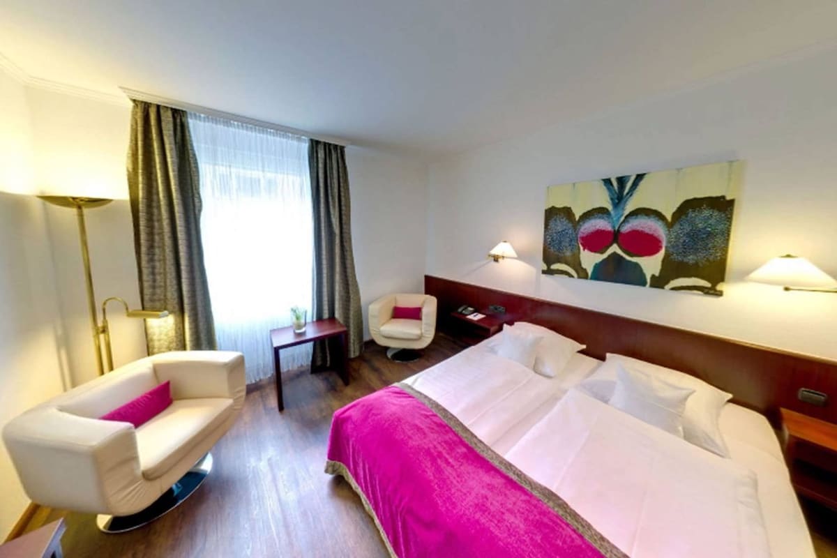 A hotel room with a bed, a chair, a desk, a lamp, and a painting on the wall.